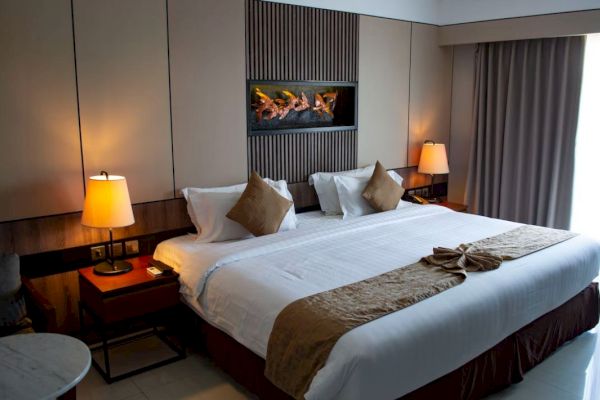 A well-lit hotel bedroom with a large bed, two side lamps, pillows, folded towels, and a fireplace décor above the bed.