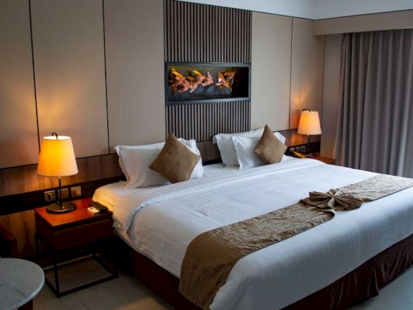 A well-lit hotel bedroom with a large bed, two side lamps, pillows, folded towels, and a fireplace décor above the bed.