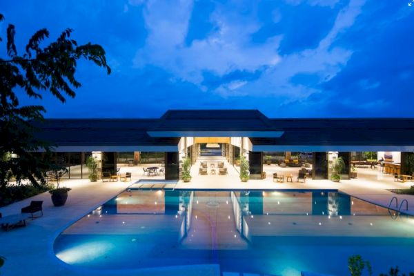 An illuminated luxury resort or hotel with a large swimming pool, surrounded by lounge chairs, against a twilight sky.