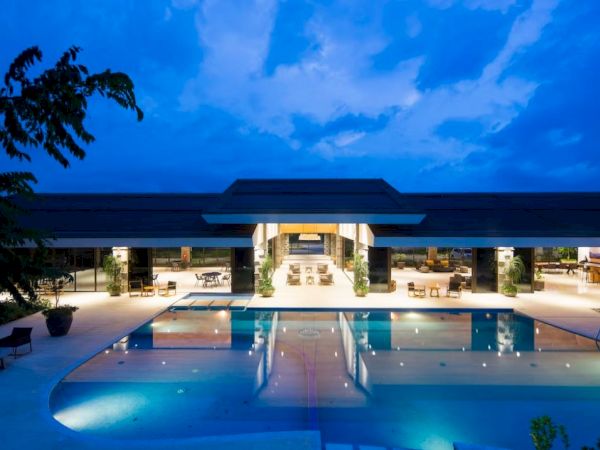 An illuminated luxury resort or hotel with a large swimming pool, surrounded by lounge chairs, against a twilight sky.