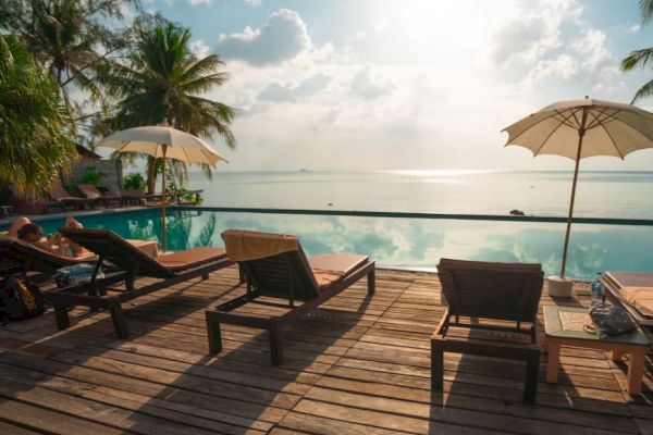 A serene poolside scene with lounge chairs, umbrellas, palm trees, and a view of a tranquil body of water under a partly cloudy sky.