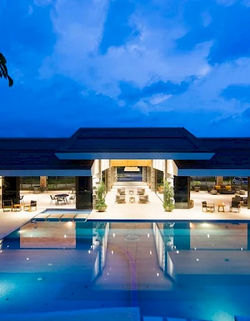 A modern, illuminated building with an inviting outdoor pool under a twilight sky, reflecting light in the water, surrounded by seating areas.