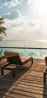 The image depicts a tranquil beach scene with lounge chairs, umbrellas, and a wooden deck overlooking a serene, reflective body of water under a bright sky.