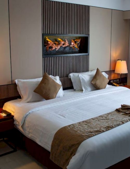 A cozy hotel room features a large bed with pillows, bedside lamps, a fireplace/TV above the bed, modern decor, and blackout curtains.