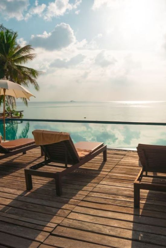 A serene poolside view with lounge chairs, umbrellas, and palm trees, overlooking a tranquil body of water under a partly cloudy sky and bright sun.