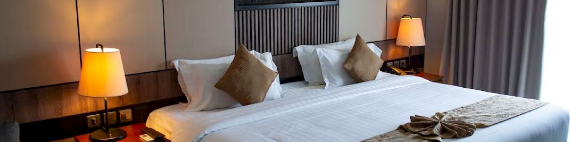 A well-lit hotel room features a large bed with white linens and brown accents, two bedside tables with lamps, and a modern fireplace.