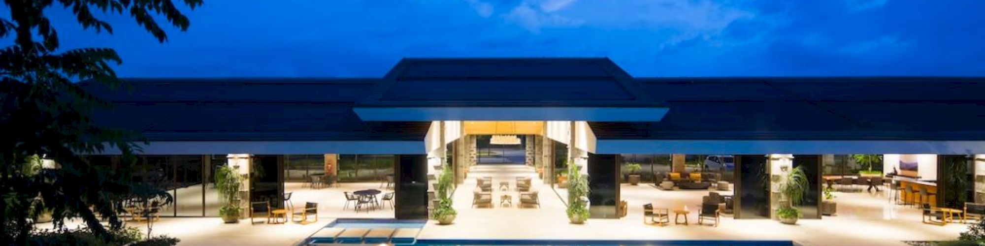 A luxurious outdoor pool area at night, with modern architecture, lounge chairs, and well-lit indoor spaces, under a partly cloudy sky.