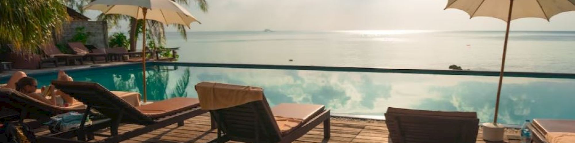 Lounge chairs and parasols on a wooden deck by an infinity pool, overlooking the ocean with palm trees and a bright, sunny sky in the background.