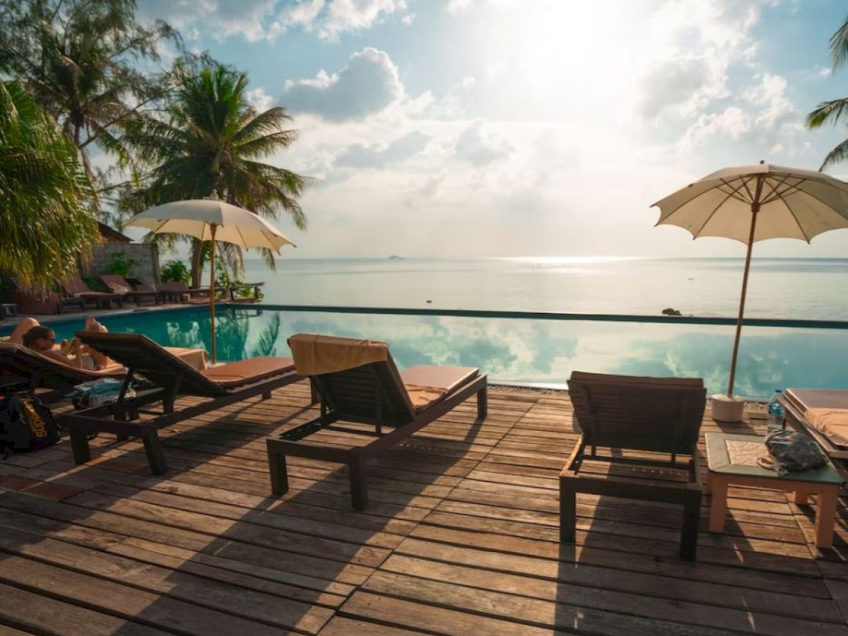 A serene poolside view with sun loungers, umbrellas, and palm trees overlooking a calm body of water under a bright sky.