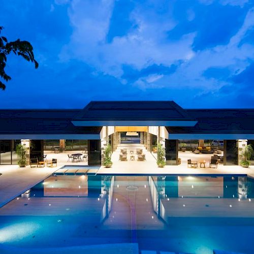 The image shows a modern building with an illuminated outdoor swimming pool on a clear evening, surrounded by lounge chairs and tables.