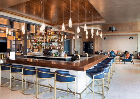 This is an image of a modern bar with a wooden counter, blue barstools, hanging lights, and a well-stocked shelf of liquor bottles.