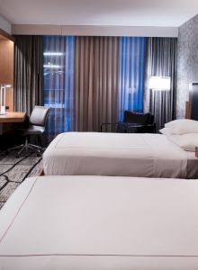 A modern hotel room with two neatly made beds, a desk with a chair, a TV on the wall, and a patterned carpet and wallpaper.