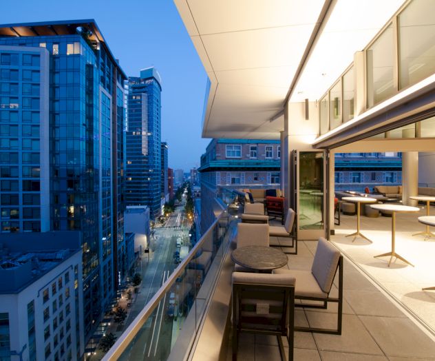 The image shows a modern cityscape viewed from a high-rise balcony at dusk, featuring a stylish outdoor seating area overlooking the city.