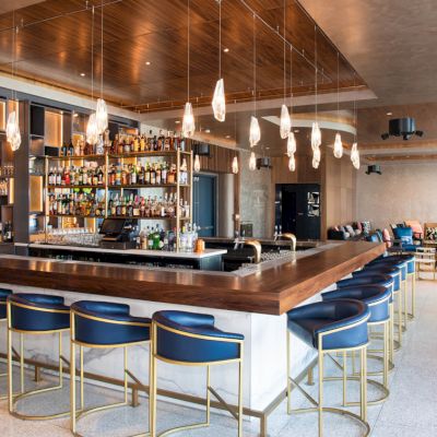 The image shows a modern, well-lit bar area with blue bar stools, a variety of bottles on display, and stylish hanging lights.