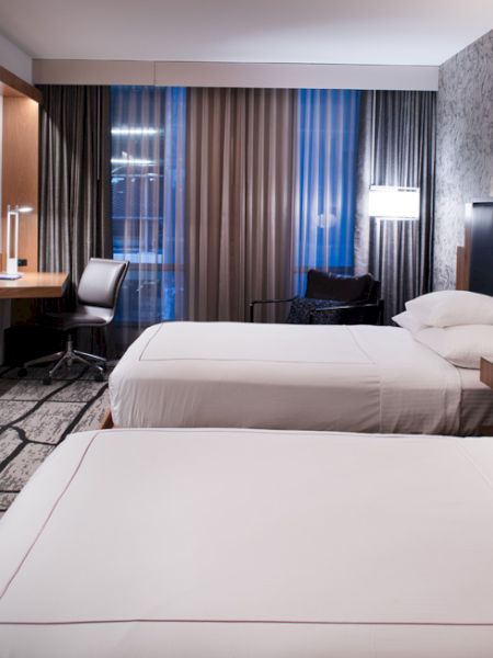 A modern hotel room with two beds, a flat-screen TV, a desk, and a large window with curtains.