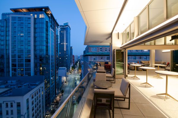 A modern cityscape view from a balcony at dusk with seating, tables, and open sliding doors leading to an indoor area with more seating, creating an inviting ambiance.
