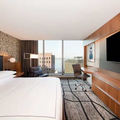 This image shows a modern hotel room with a large bed, TV, desk, and a window with a city view. The decor includes wood paneling and patterned walls.