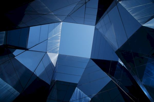 The image shows a geometric architectural structure with reflective glass panels, forming an open square revealing a clear blue sky at the center.