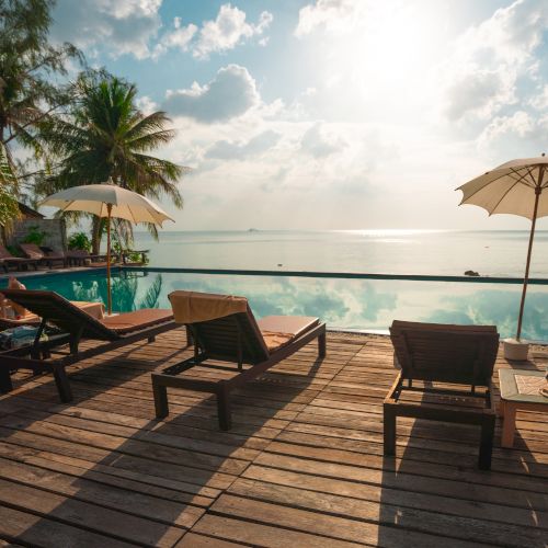 Poolside wooden deck with lounge chairs and umbrellas, overlooking a calm sea under a partly cloudy sky, with lush greenery around.