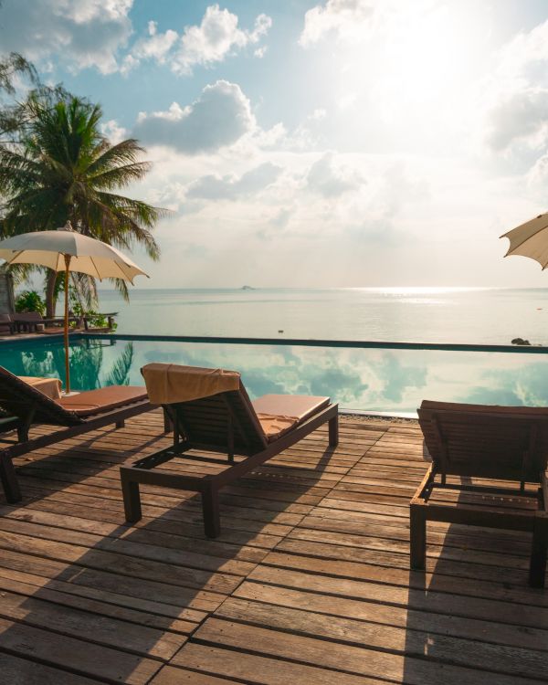 Poolside wooden deck with lounge chairs and umbrellas, overlooking a calm sea under a partly cloudy sky, with lush greenery around.