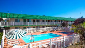 This image shows a motel with a green roof, featuring a fenced outdoor pool area with lounge chairs, tables, and a green-striped umbrella by the pool.