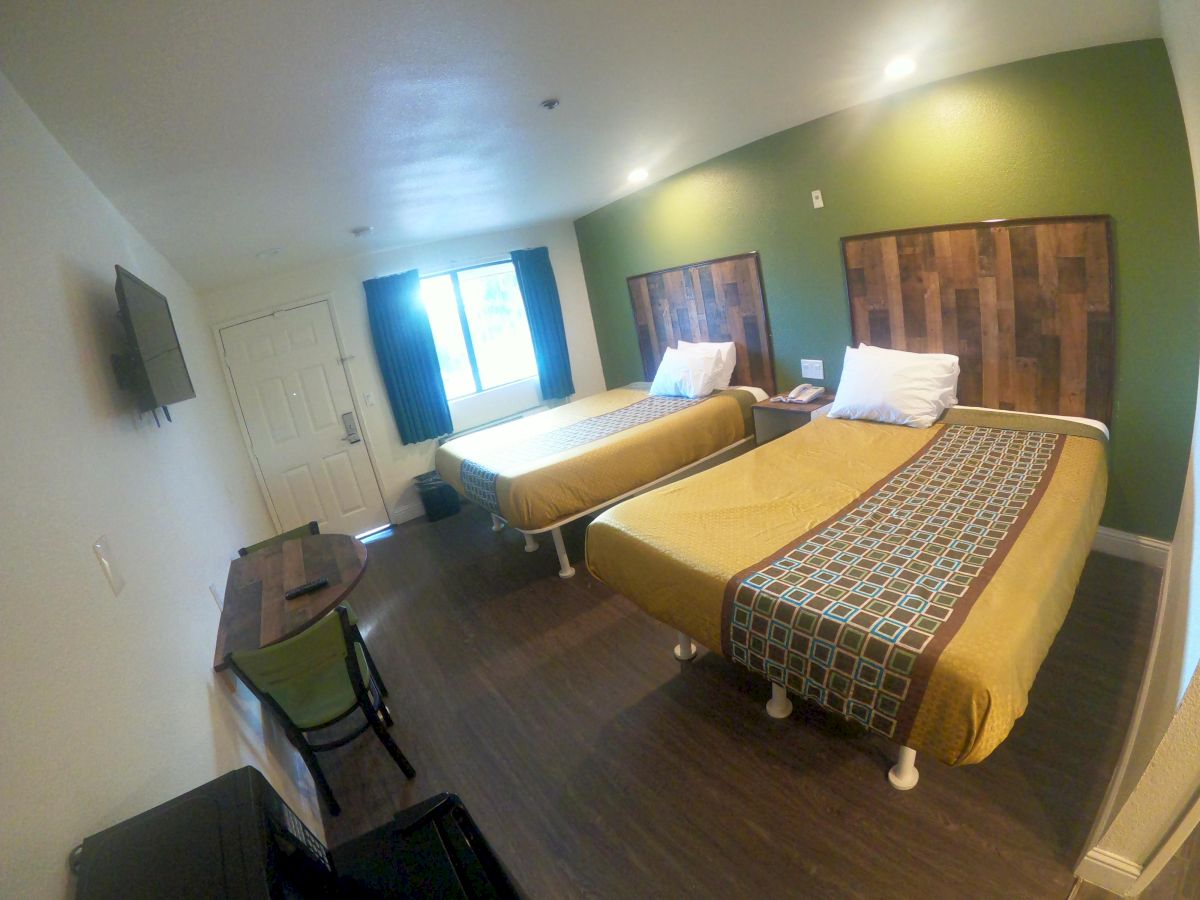 The image shows a hotel room with two beds, green-accented walls, a wall-mounted TV, a desk with a chair, and a window with curtains.