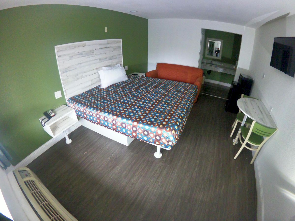 A hotel room with a bed, an orange sofa, a wall-mounted TV, a small table with chairs, and a mirror. The bed has a colorful patterned cover.