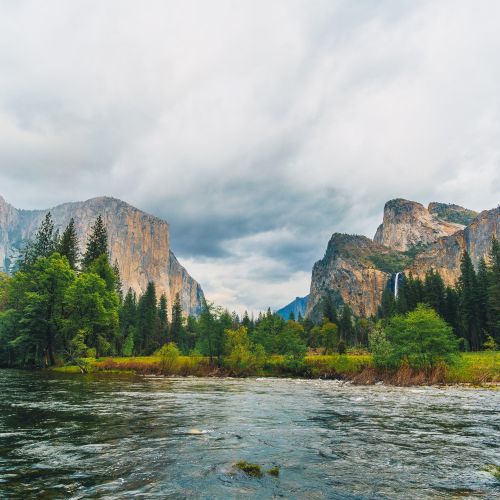 The image shows a scenic view of a river surrounded by lush greenery and tall, rugged mountains with cloudy skies overhead, creating a serene landscape.