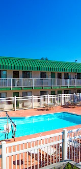 The image shows an outdoor pool area with white fencing, green-roofed buildings, and lounge chairs under striped umbrellas under a clear blue sky.