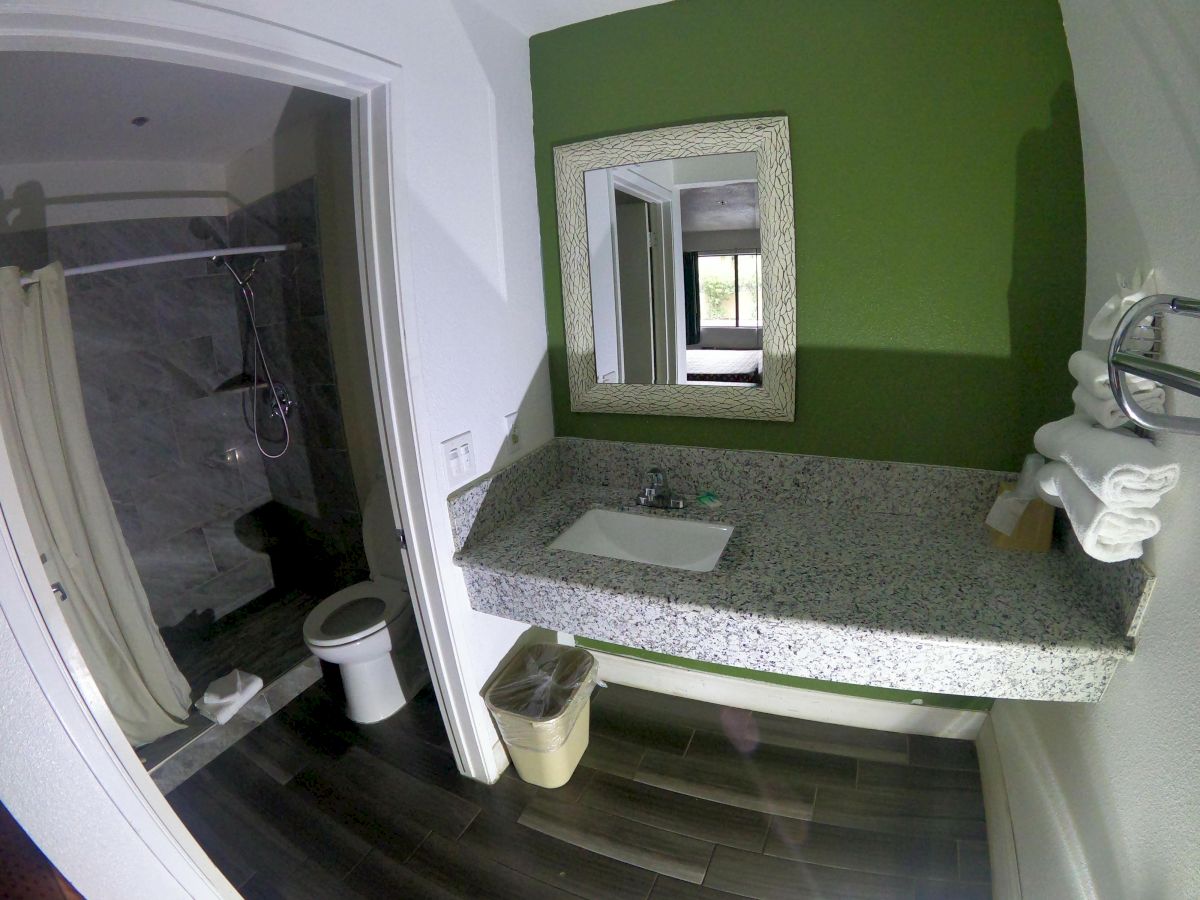 A bathroom with a green wall, a sink, a mirror, a trash can, towels, a toilet, and a shower area with a curtain is shown.