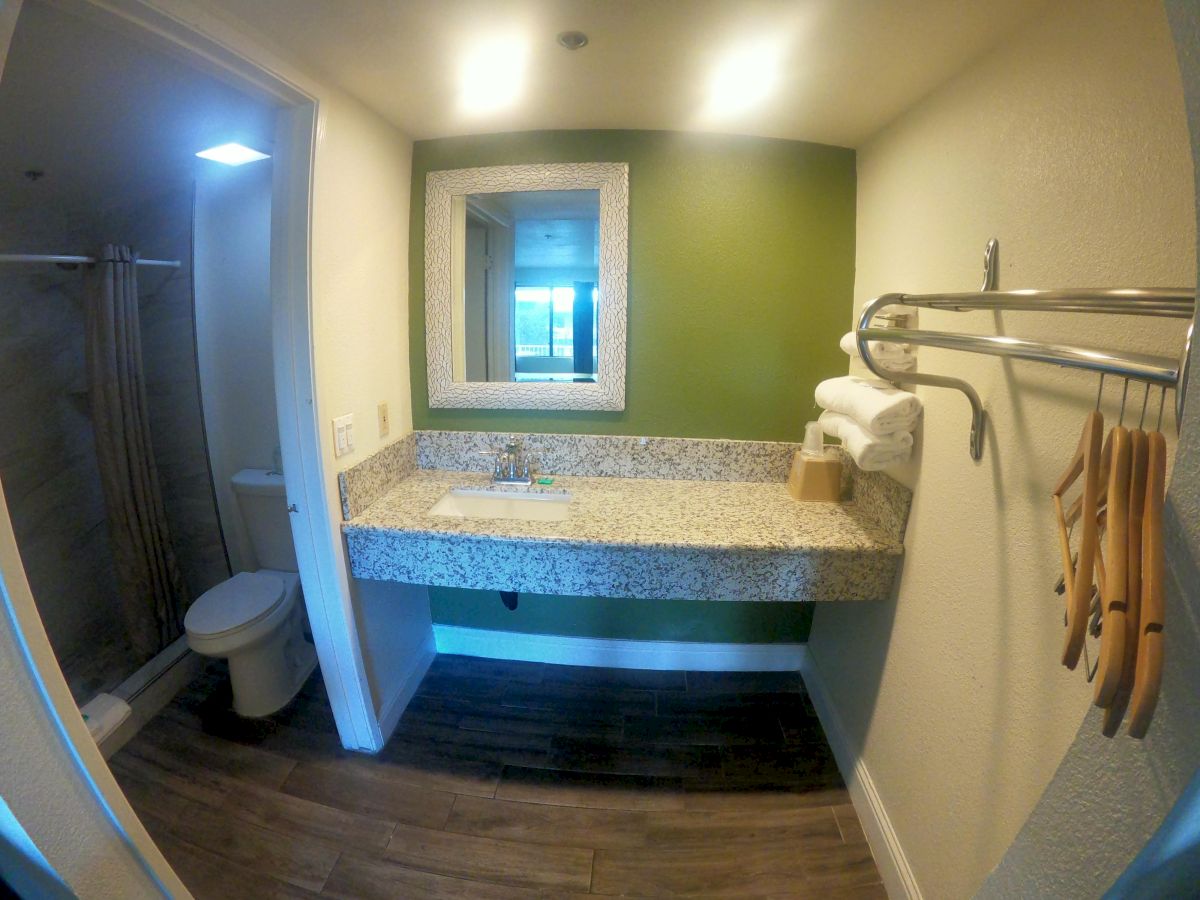 A bathroom with a sink and mirror, towels on a rack, wooden hangers, and a toilet and shower in a separate area, with light shining from above.