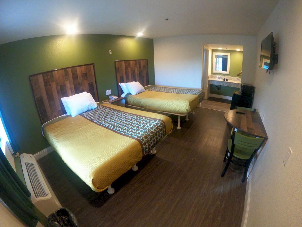 A hotel room with two beds, a table and chairs, a TV on the wall, and a sink area with a mirror visible in the background, under 140 characters.