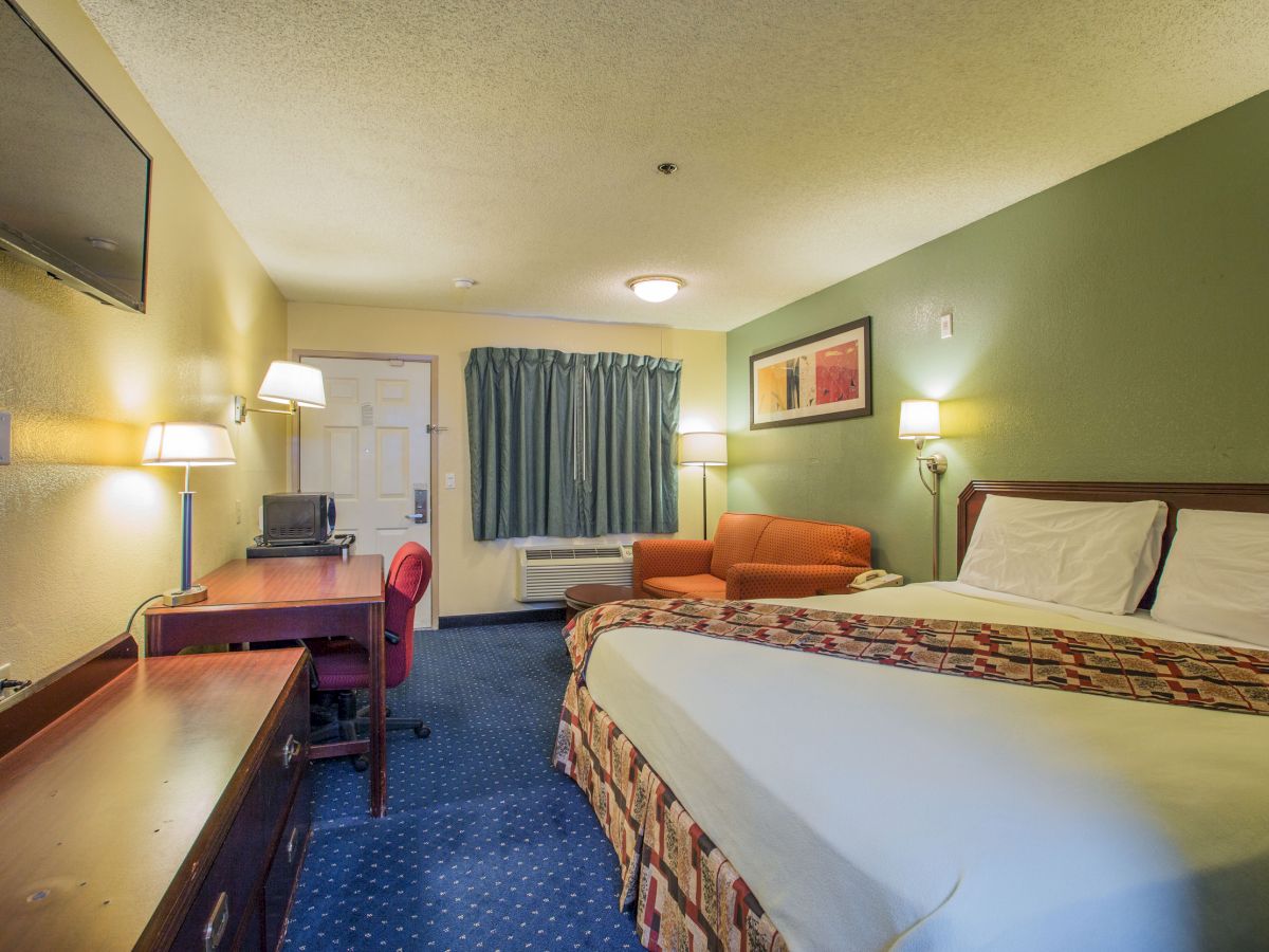 A hotel room with a double bed, desk, chair, sofa, TV, green accent wall, and a window with blue curtains.