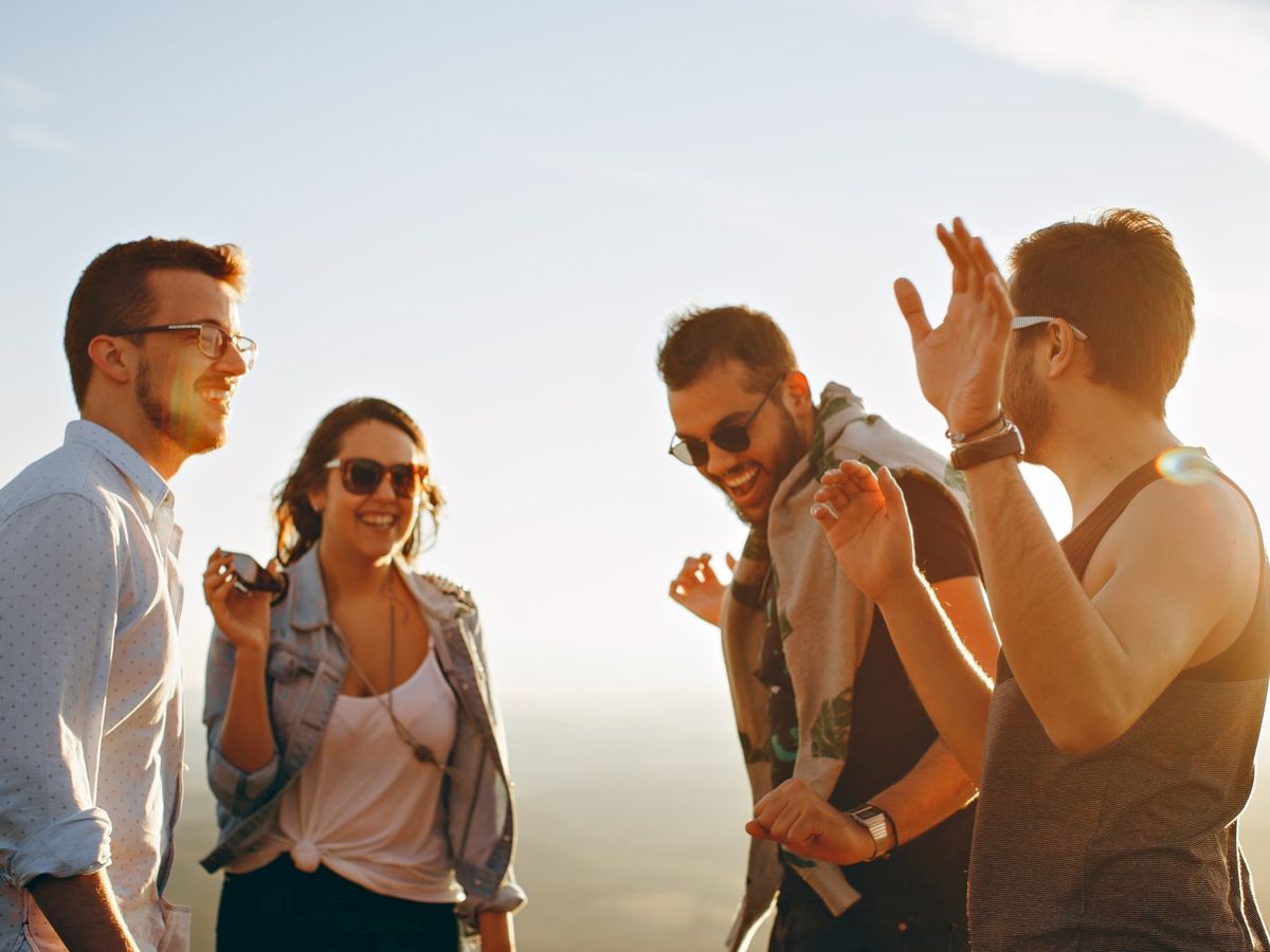 A group of four people, two men and two women, are outdoors in the sunlight. They are smiling, laughing, and appear to be enjoying each other's company.