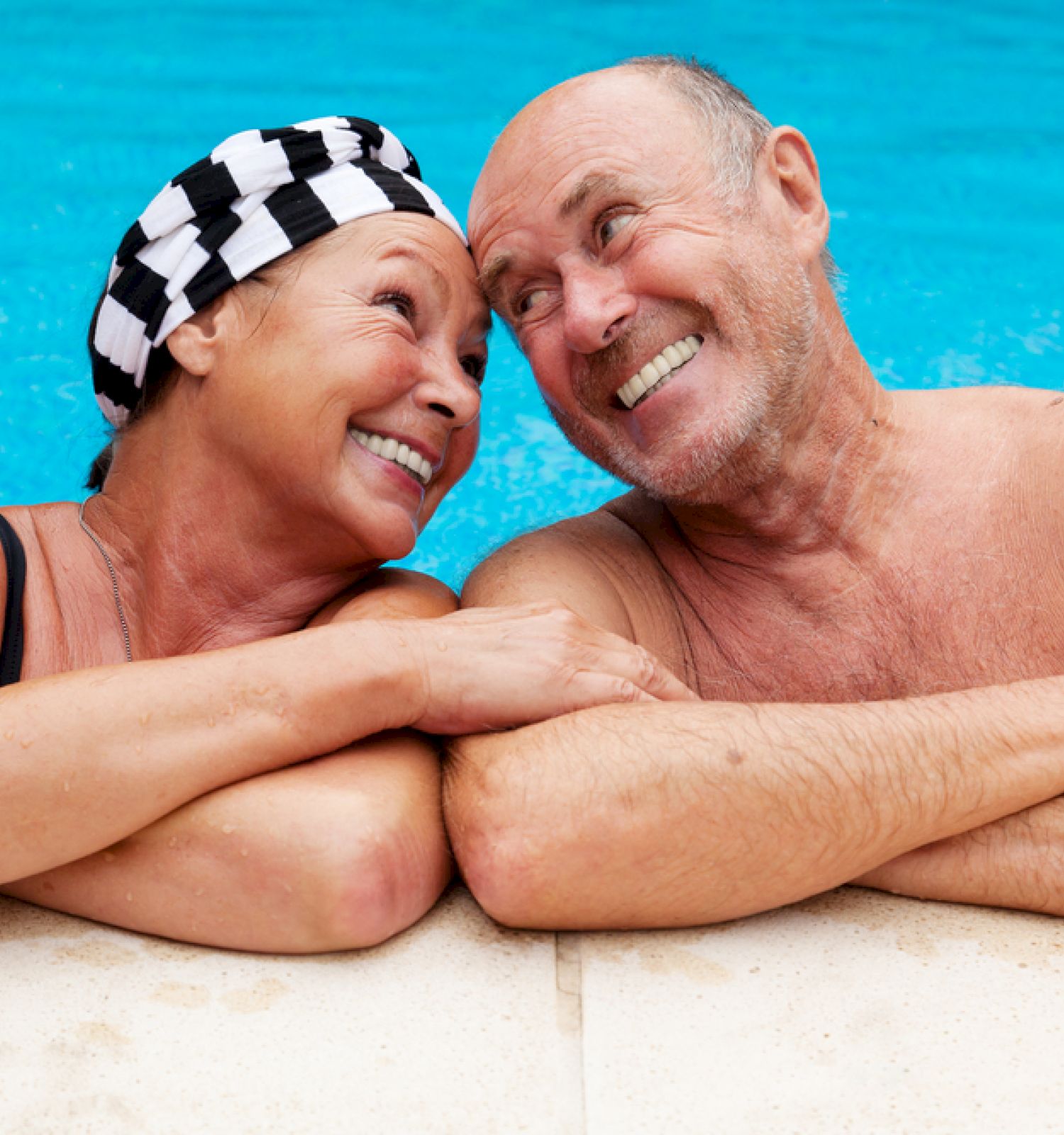 An elderly couple is smiling and relaxing by the edge of a swimming pool, with water in the background.