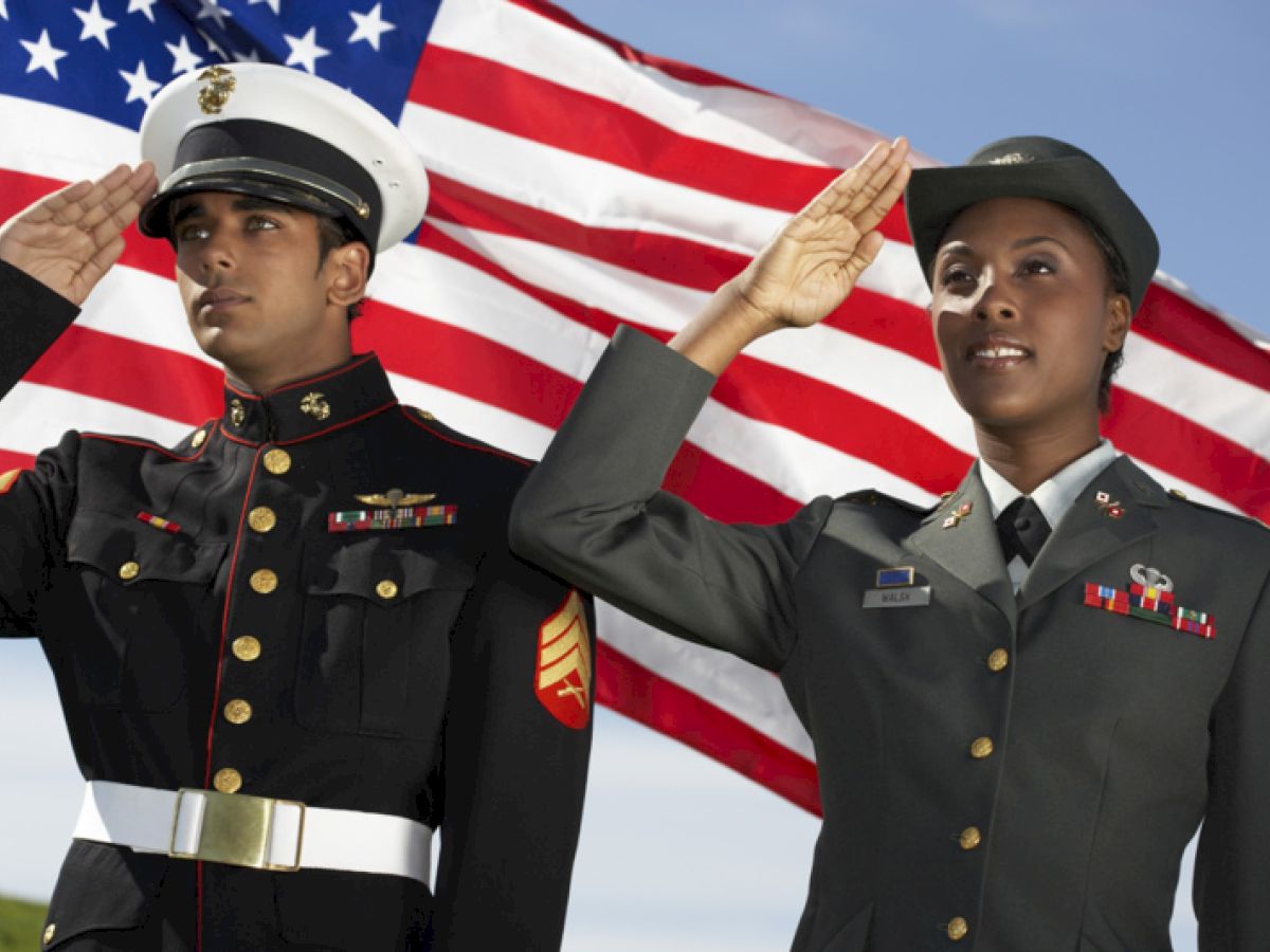 Two military personnel are saluting in front of an American flag, one in a marine uniform, the other in an army uniform.