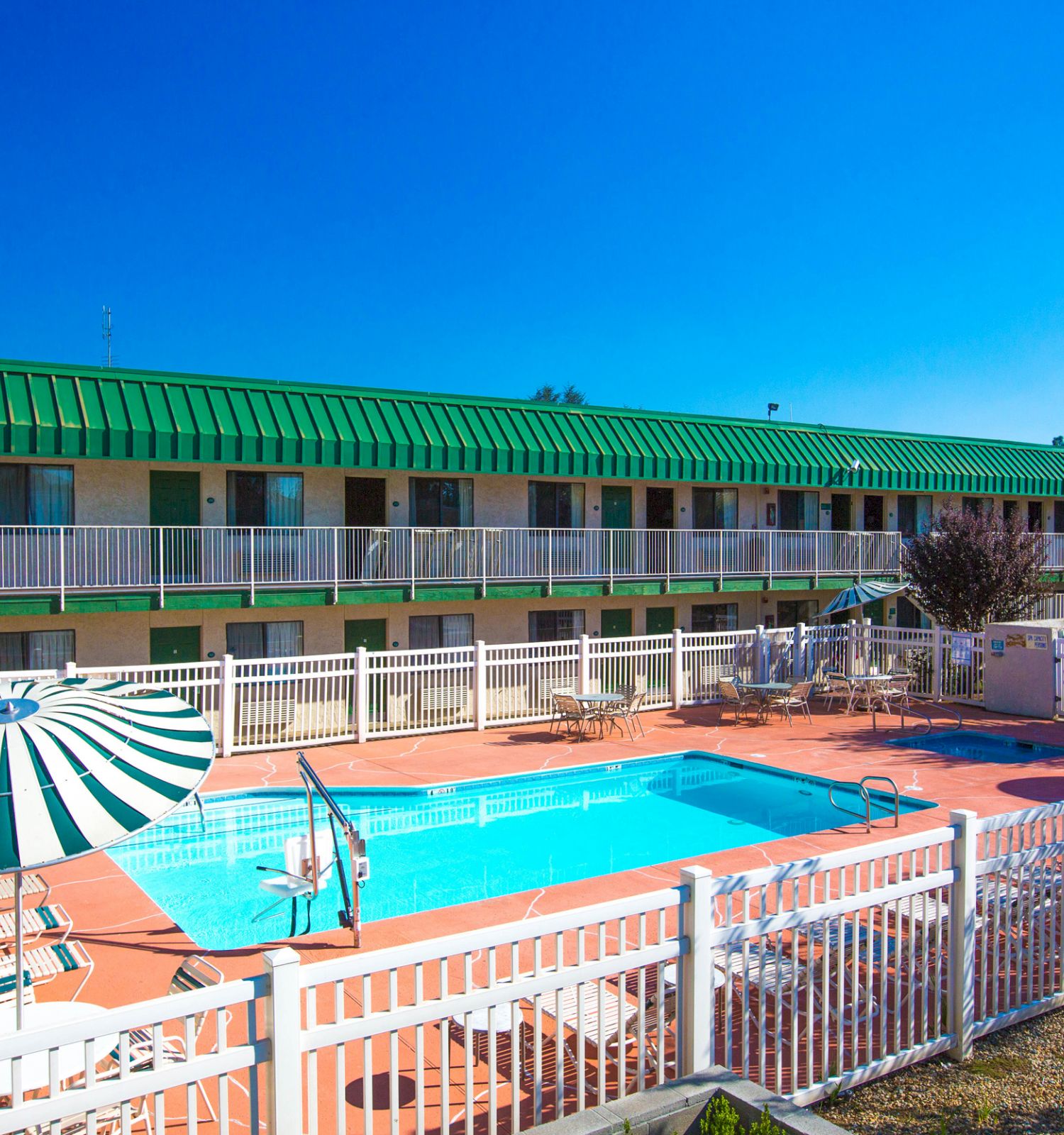 This image shows an outdoor swimming pool area with lounge chairs, umbrellas, and a two-story building with green roofs in the background.