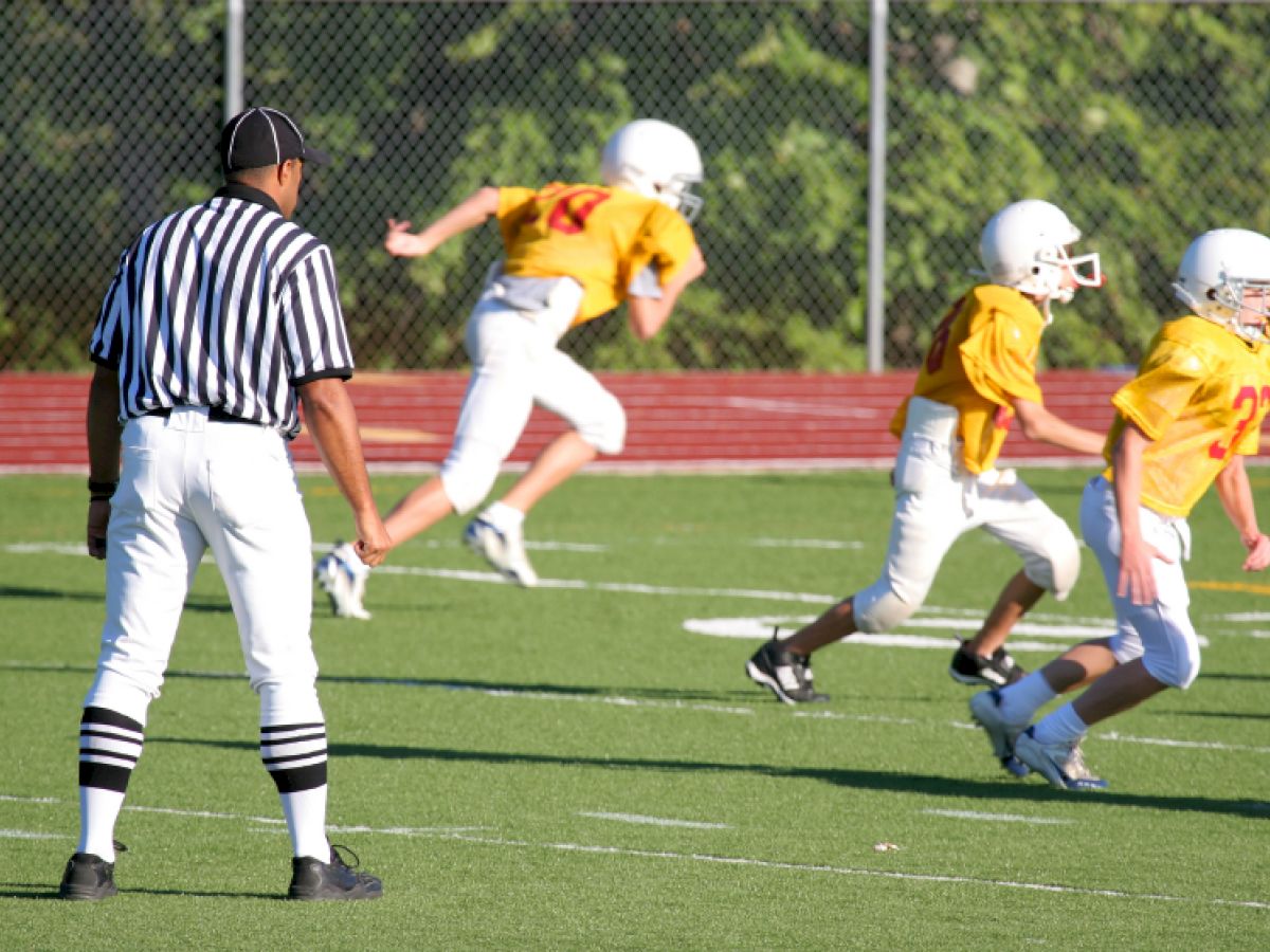 A referee oversees a youth football game as players in yellow jerseys and white helmets run on the field, surrounded by a grassy background.