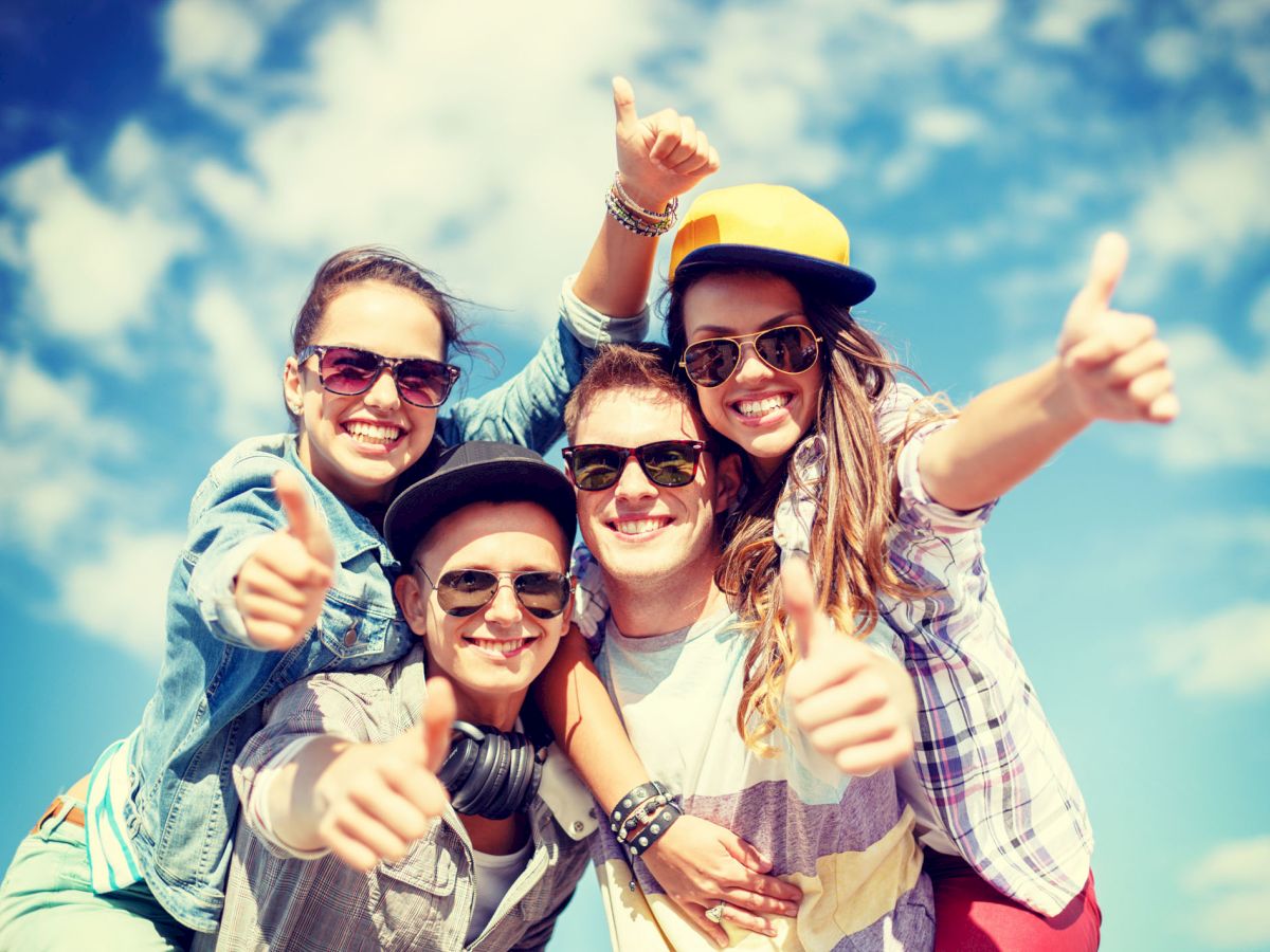 A group of four friends, wearing sunglasses and casual clothing, smile and give thumbs up against a bright blue sky with some white clouds.