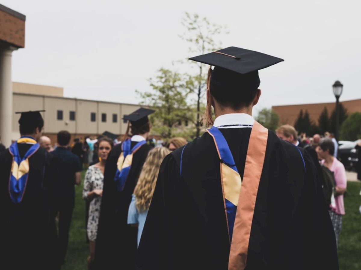 Graduates in caps and gowns gather with people on a lawn near buildings, likely at a graduation ceremony, on a somewhat cloudy day.