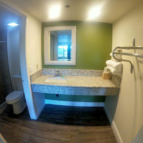 A bathroom with a green accent wall, granite countertop, mirror, towels on a rack, a few hangers, and a shower and toilet visible in the adjacent room.