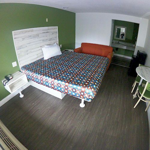 A modern hotel room features a colorful bed, an orange couch, a wall-mounted TV, a small desk with a chair, and a green accent wall.