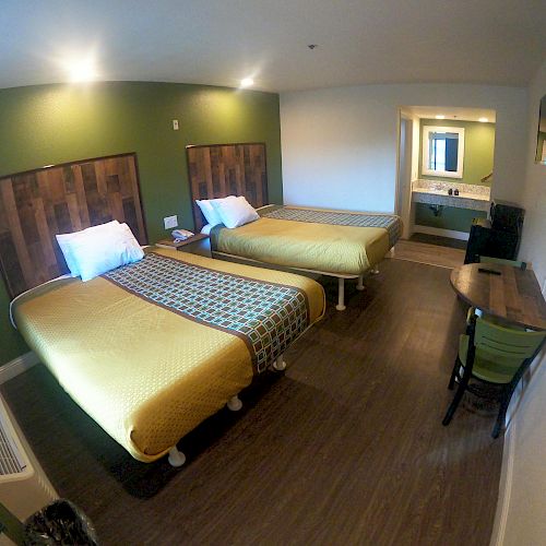 The image shows a hotel room with two beds, a table with chairs, wall-mounted TV, and a vanity with a sink. The decor is modern and minimalistic.