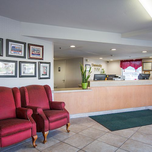 The image shows a reception area with a front desk, red armchairs, framed pictures on the wall, potted plants, and a green rug near the entrance.