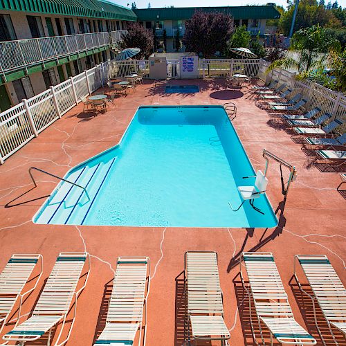 An outdoor pool area with lounge chairs arranged around it, surrounded by a fence and adjacent to a two-story building.