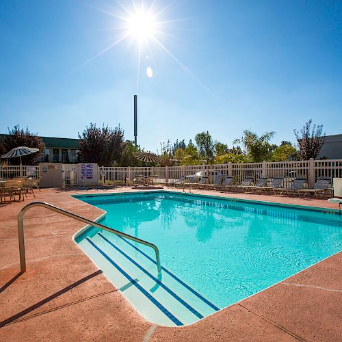 An outdoor swimming pool with clear blue water, surrounded by a fenced area and lounge chairs, under a bright sunny sky.