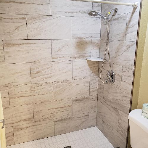 A bathroom with a tiled shower area, a showerhead, a small shelf, a toilet, and a folded towel on the floor near the toilet.