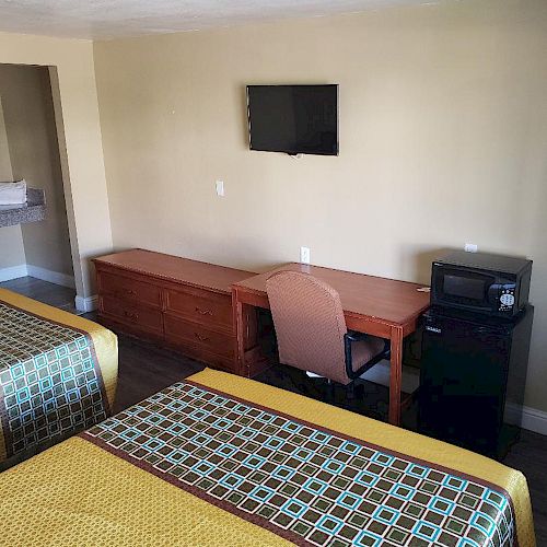 A hotel room with two beds, a small desk with a chair, a wall-mounted TV, and a microwave on a mini-fridge. The room has beige walls.