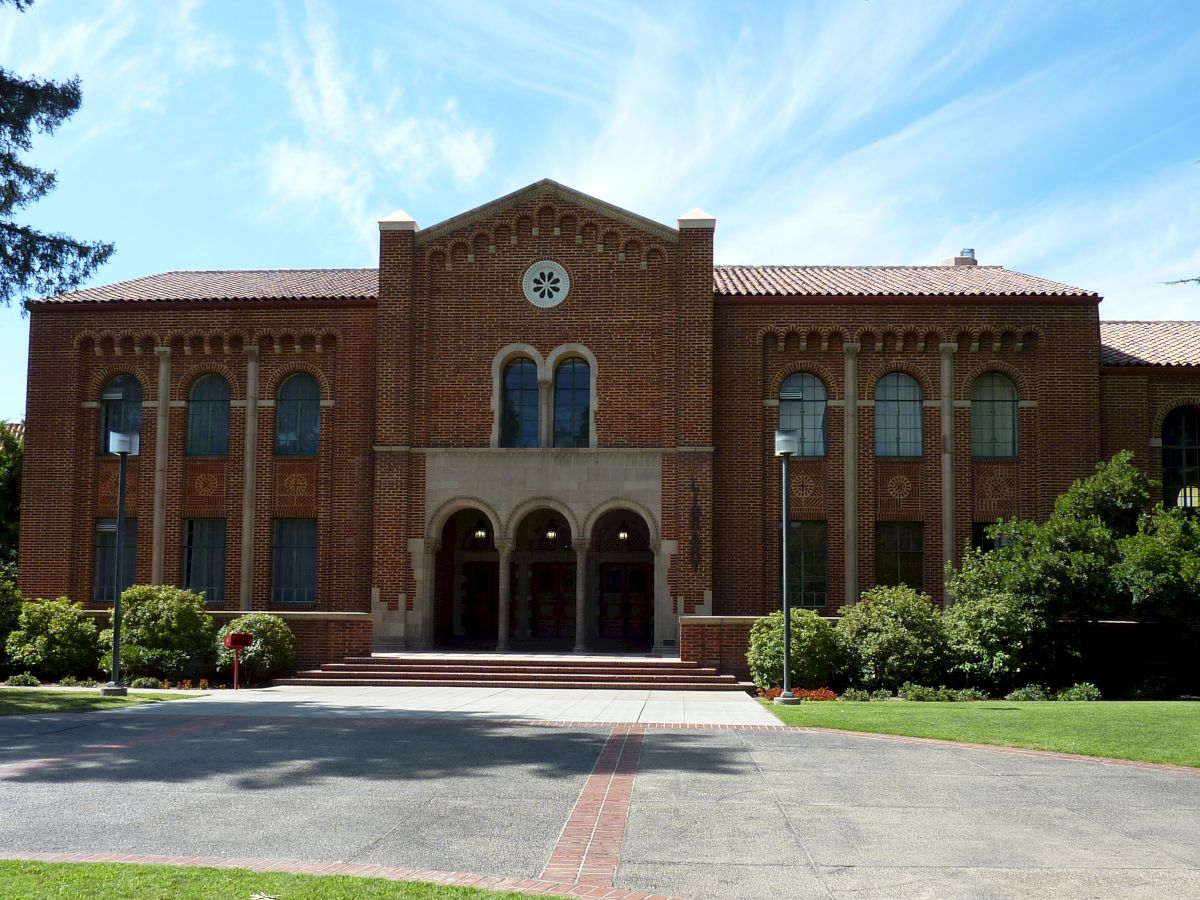 The image shows a large brick building with a clock at the top center and an arched entryway, surrounded by greenery and trees.