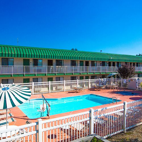 The image shows an outdoor swimming pool surrounded by a white fence, with lounge chairs, umbrellas, and a two-story building in the background.